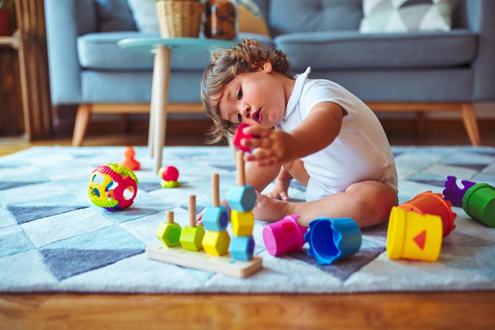 Um bebê está sentado no tapete de uma sala de estar mexendo com brinquedos de empilhar e montar, que é uma das brincadeiras que estimulam o desenvolvimento infantil. 
