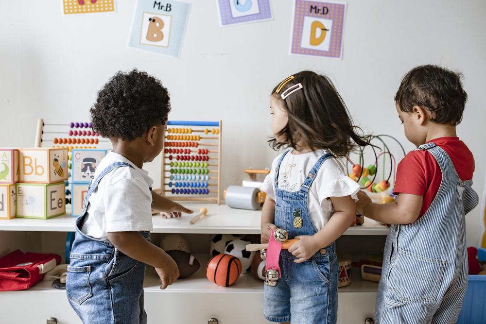 Três crianças brincam com brinquedos diversos dentro de uma sala de creche.