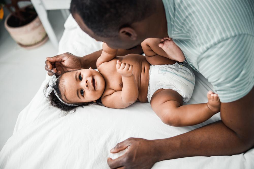 Antes do desfralde: imagem de um pai está apoiado na cama, fazendo um bebê de fralda sorrir. Ele faz carinho na cabeça da criança.  