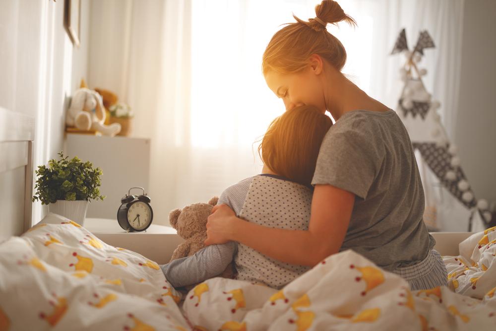 Mãe, sentada na cama, abraça o filho que acabou de acordar, representando o ensino da disciplina positiva. 