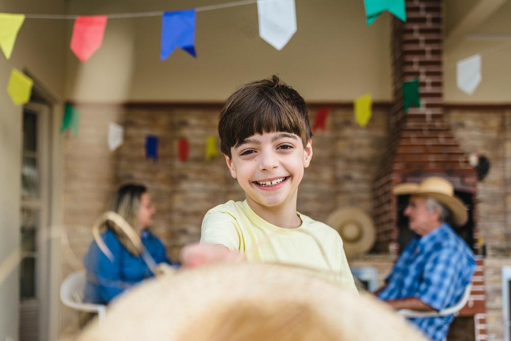 Criança sorrindo para a foto. Ao fundo, dois adultos conversam num cenário de festa junina, com bandeirinhas coloridas penduradas.