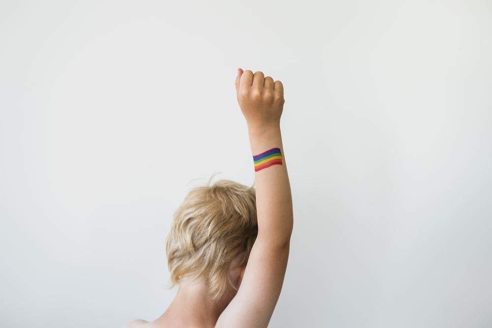 Foto de uma criança trans de costas com um braço levantado. Quase na altura de seu pulso, se vê uma pintura que traz as listras coloridas do arco-íris, um símbolo LGBTQIA+.