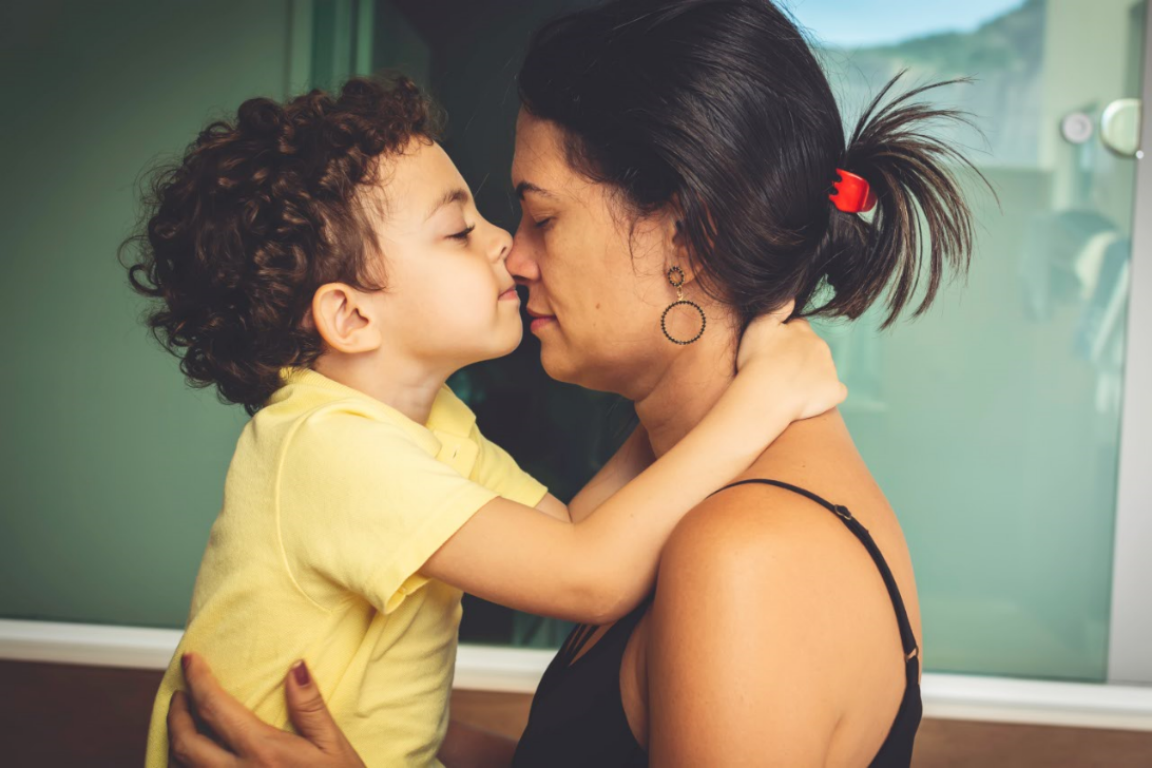 Filho e mãe, frente a frente de olhos fechados, se abraçam enquanto encostam os seus narizes. A imagem busca representar a relação com um filho que tem TDAH.