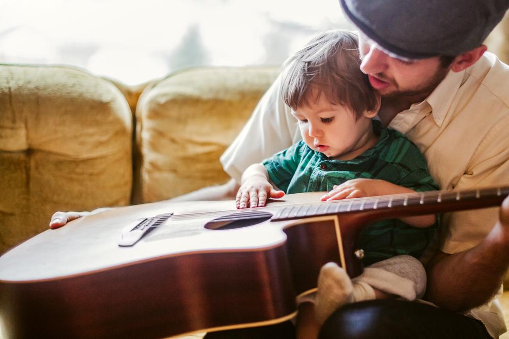 Um homem segura seu filho no colo e, juntos, eles seguram um violão, usando da música para estimular o desenvolvimento infantil.