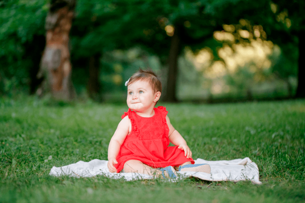 Criança de um ano com vestido vermelho e fita no cabelo posa para foto em um parque, sentado em um pequeno cobertor colocado sobre a grama.