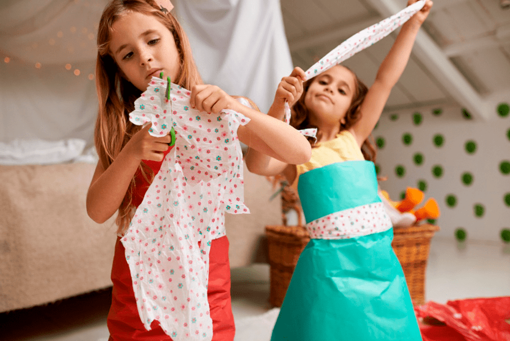 Duas crianças de cerca de 8 anos cortam papel colorido com uma tesoura sem ponta customizando pequenas peças de roupa com este material. Uma delas está utilizando, sobre a sua roupa comum, um vestido feito de papel.