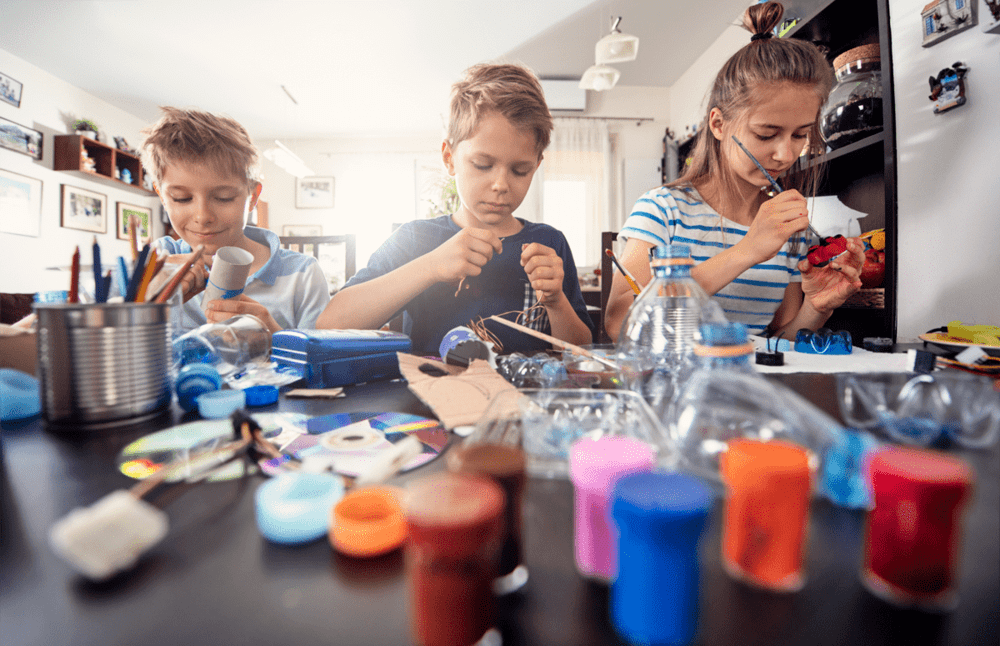 Três pré-adolescentes pintam brinquedos não estruturados sentados à mesa em uma cozinha. Esses brinquedos estão sendo criados a partir de garrafas pet cortadas, papelão e outros materiais.