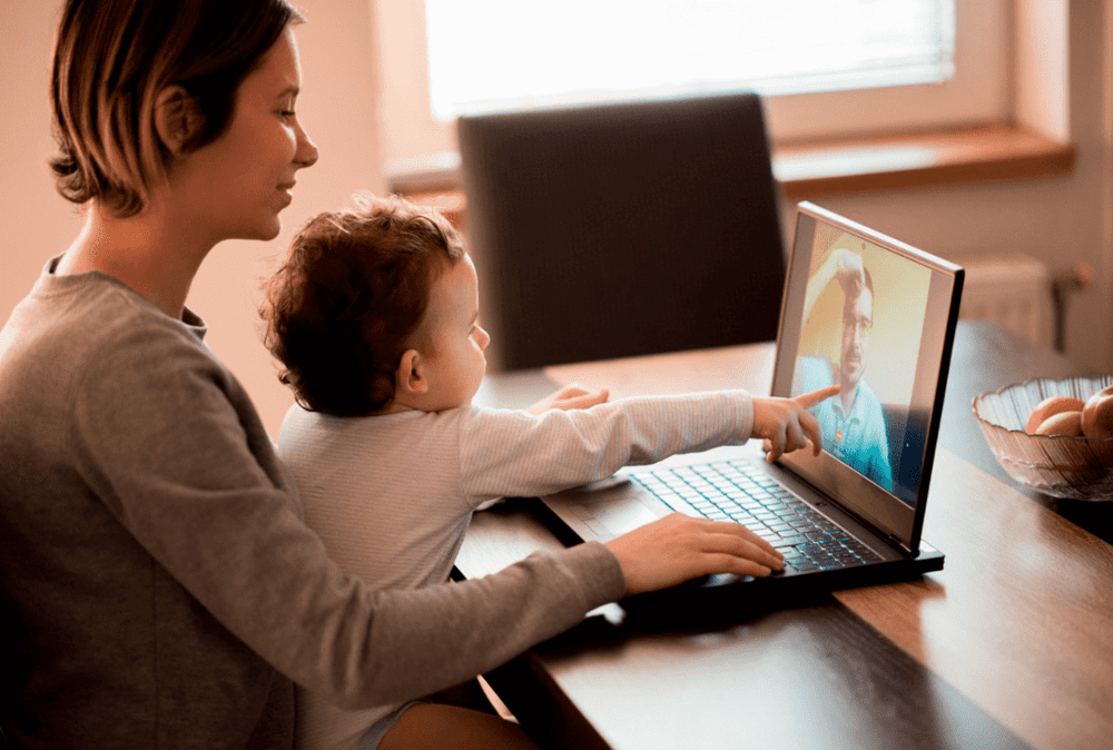 Ilustrando a separação dos pais, uma mãe segura um bebê em seu colo. Eles estão sentados à mesa em frente ao computador. O bebê aponta para a tela, onde há um homem por videochamada.