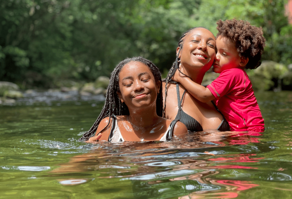 Dentro de um lago, a cantora Negra Li, ilustra a importância da música para crianças. Ela está segurando um filho no colo e em frente está sua outra filha.