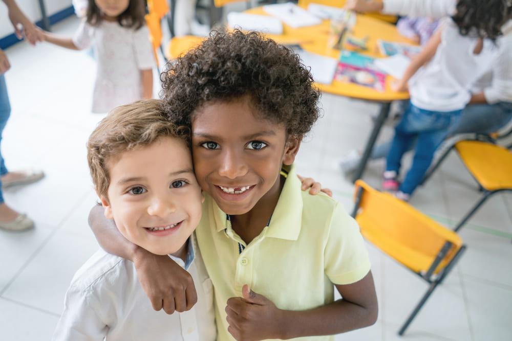 A foto mostra dois meninos de cores e aparências distintas sorrindo, se abraçando e olhando para a câmera, dentro de uma sala de aula. 