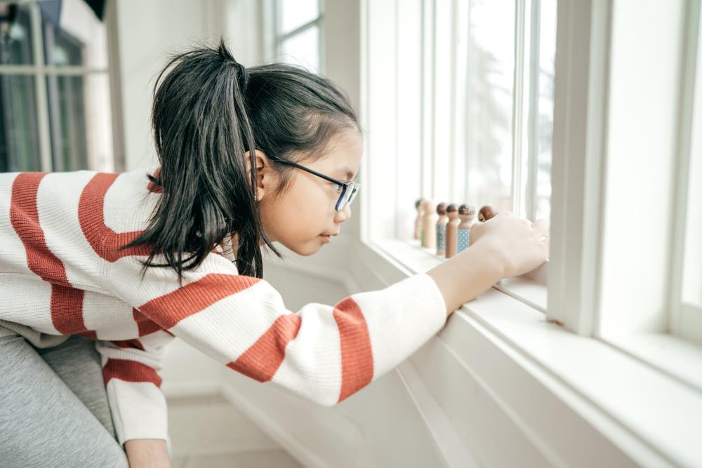 Na imagem, há uma menina vestida com blusa listrada e calça cinza, usando rabo de cavalo e óculos. Ela está agachada em frente à janela, enfileirando suas mini bonecas de madeira demonstrando perfeccionismo, uma das características do autismo 