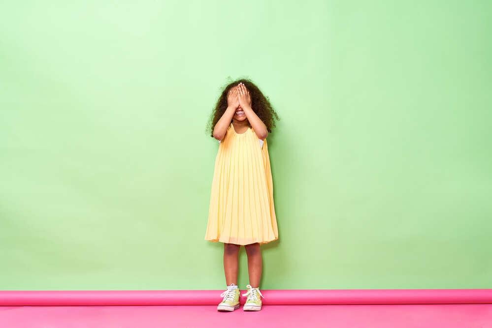 Uma criança tímida sorrindo, mas tampando o rosto com as mãos. Ela está de vestido amarelo, em frente a uma parede verde.