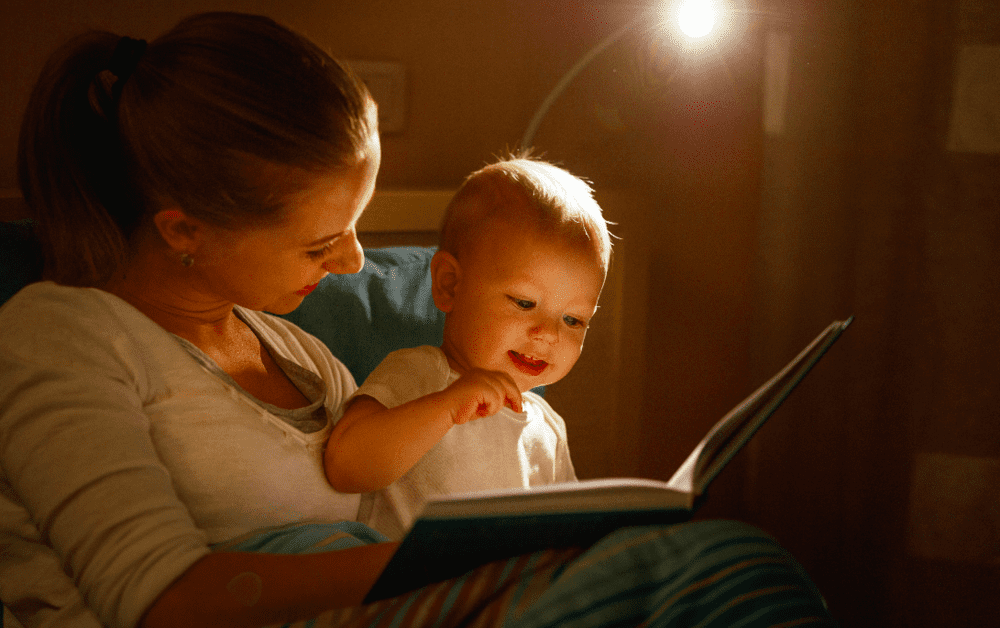 A mãe sorri com o filho bebê no colo enquanto conta uma história para dormir em um livro para ele.