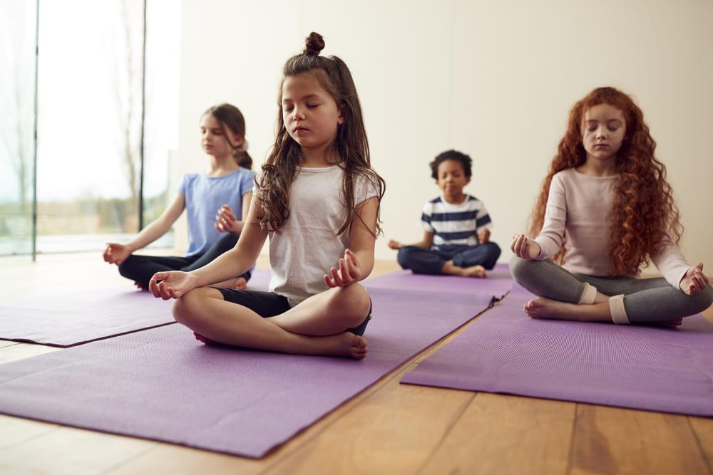 A imagem mostra quatro crianças sentadas praticando meditação infantil, com as pernas cruzadas e os olhos fechados.