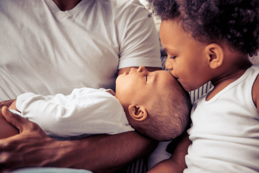 Uma criança beija seu irmão no topo da cabeça, na região da moleira, enquanto o bebê dorme no colo do pai.