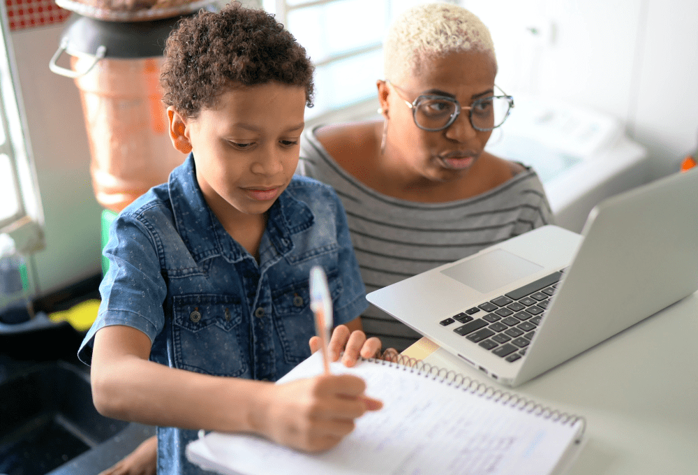 Mãe auxilia filho durante aula on-line na pandemia, enquanto ele escreve em um caderno.