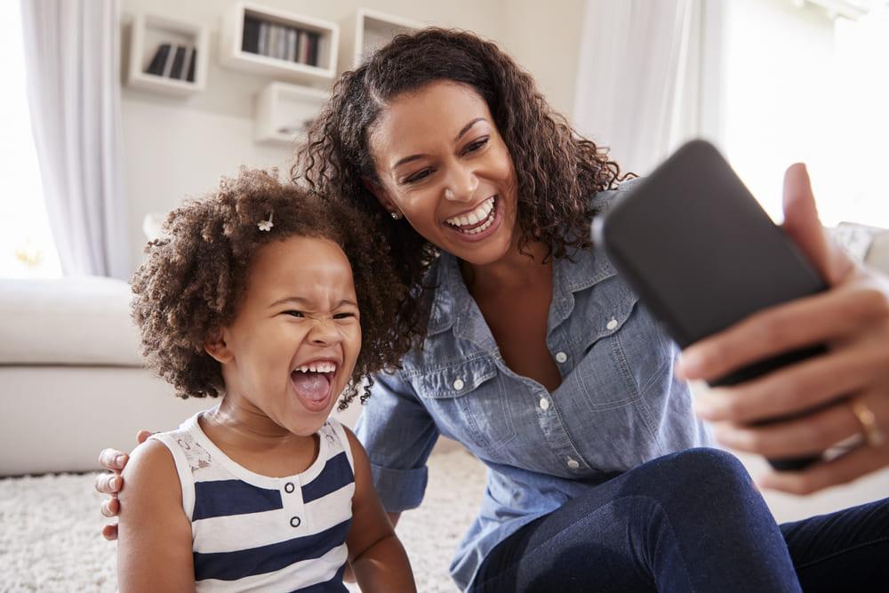 Sentadas no chão, mãe e filha sorriem ao tirar uma foto com o celular. As duas apresentam uma personalidade parecida.