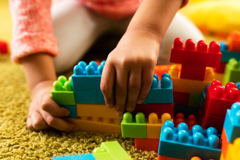 A imagem mostra as mãos de uma criança montando peças de brinquedo em cima de um tapete, representando a importância do brincar