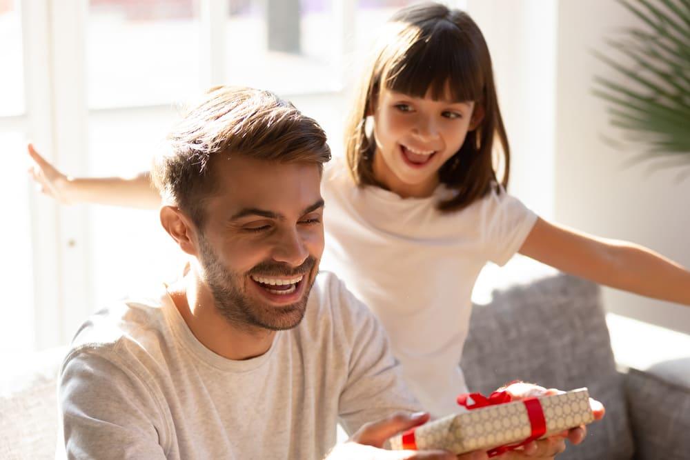 Pai e filha sorrindo enquanto o pai segura um presente de dia dos pais, ambos estão em uma sala bastante iluminada.