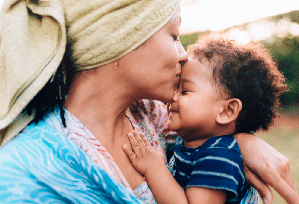 Mãe tem uma toalha de banho enrolada no cabelo e beija a testa do filho, eles estão de olhos fechados, como se satisfeitos, ilustrando a importância da rede de apoio.