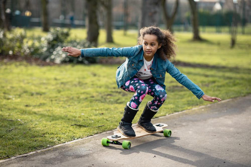 Foto de uma menina andando de skate ao ar livre, em um lugar plano. Um incentivo a pratica de skate infantil.