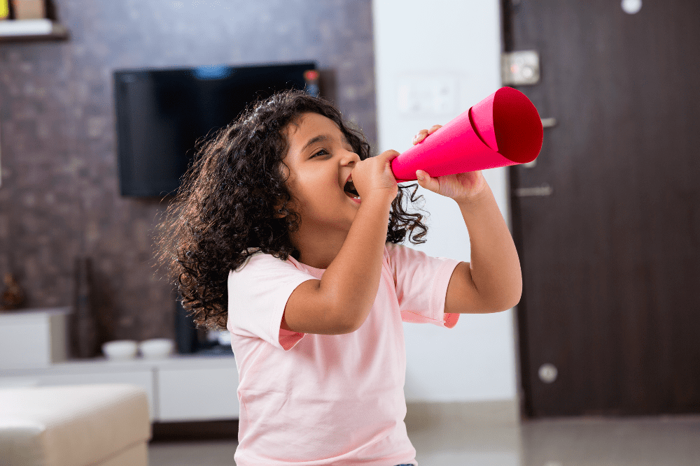 Menina sorri enquanto grita algo utilizando um megafone feito de papel enrolado, ilustrando a hiperatividade.