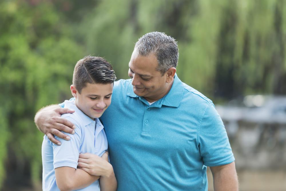 Pai e filho, ambos vestidos de azul, se abraçam em um ambiente aberto mostrando o vínculo emocional entre pai e filho