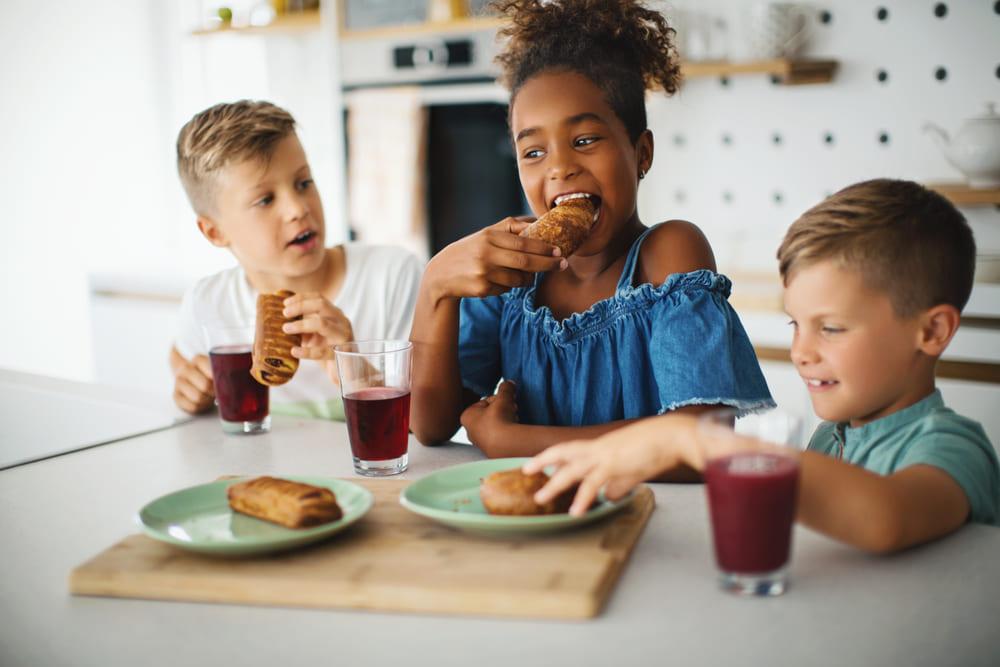 Na imagem, 3 estão crianças sentadas em frente à uma mesa de cozinha. A menina está no meio e os dois meninos estão nos cantos. Eles estão tomando suco e comendo sanduíches