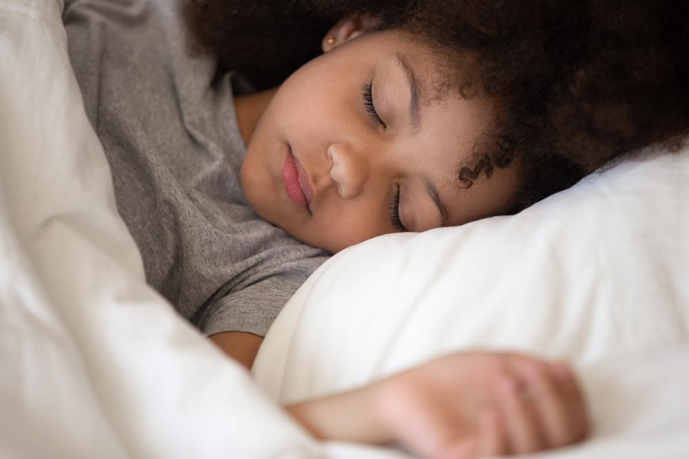 Menina dorme sobre uma cama, com a cabeça apoiada no travesseiro. A imagem ilustra bons hábitos, como o sono regulado.
