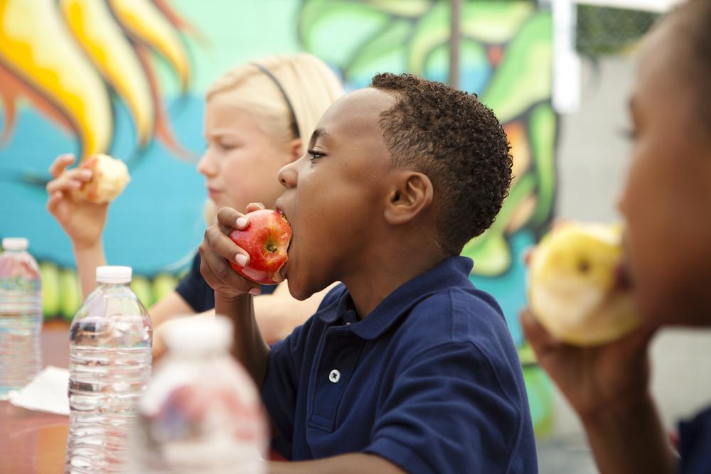 A imagem mostra três crianças vestindo uniforme azul, sentadas e comendo maçã (fonte de nutrientes para crianças). Na mesa, há 3 garrafas de plástico com água.  
