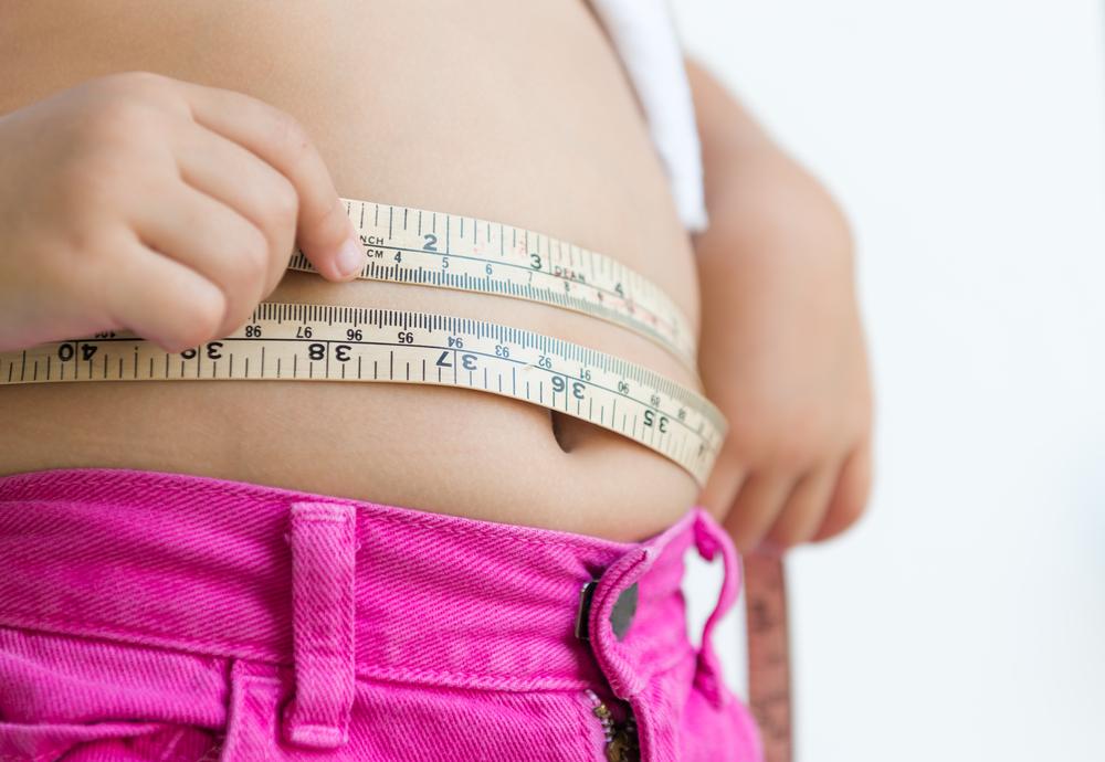 Representando a obesidade infantil, a imagem mostra a região da barriga de uma criança enquanto ela mede sua curvatura com uma fita métrica.