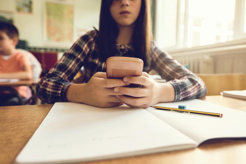 Representando o vício em redes sociais, menina mexe no celular, com os braços apoiados sobre uma mesa com um caderno