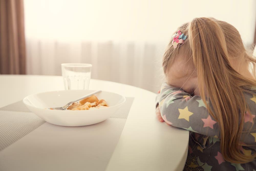 Menina se recusa a comer. Ela está escorada na mesa com a cabeça baixa e braços cruzados. À frente dela tem um prato de comida e um copo. 