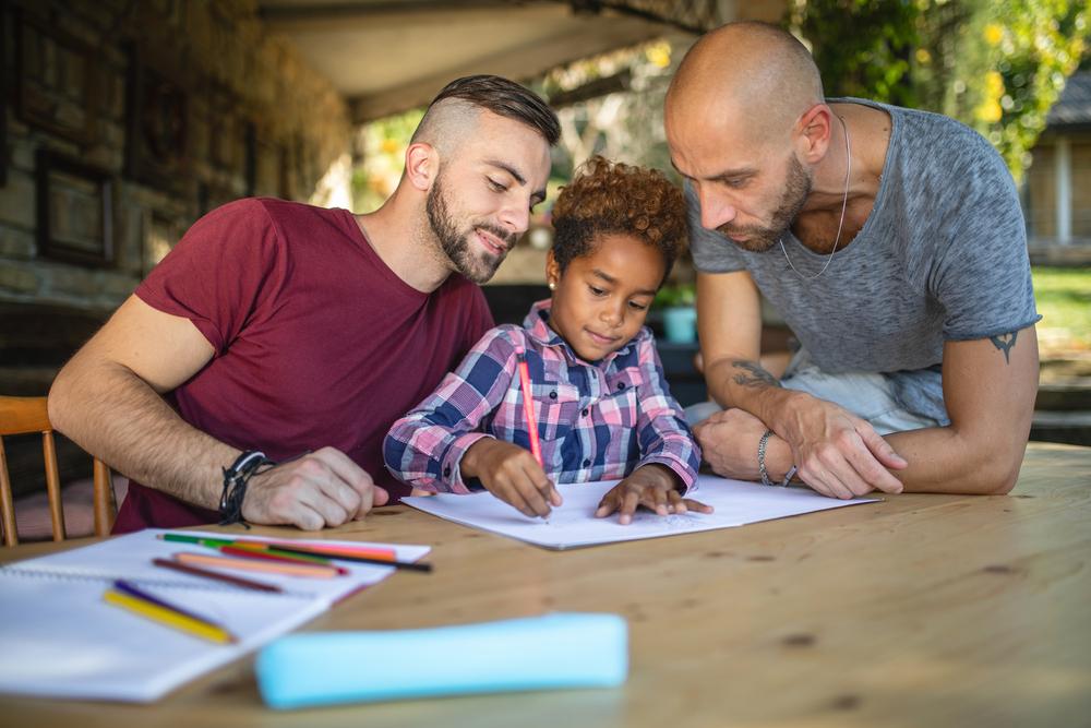 Dois homens observam sorrindo a filha desenhar em uma folha de papel, em uma mesa com lápis de cor espalhados e papéis.