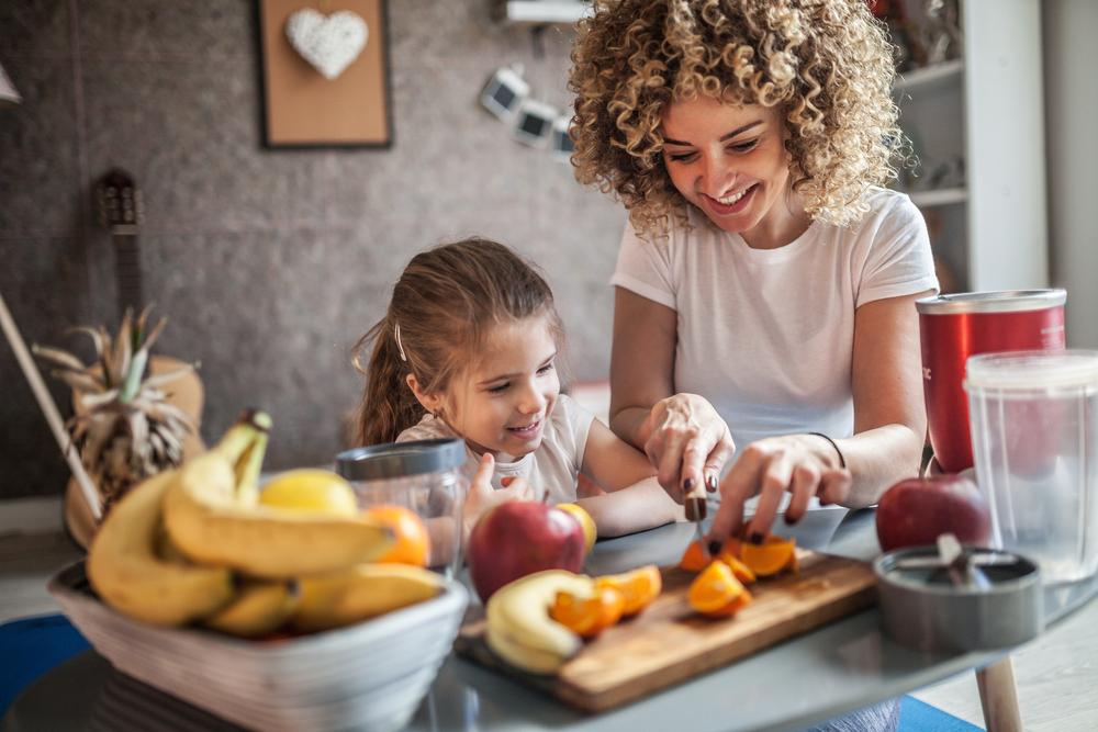 Mãe corta uma fruta para preparar um café da manhã infantil, enquanto a filha observa. Ambas sentadas no chão e apoiadas em uma mesa com diversas opções de frutas.