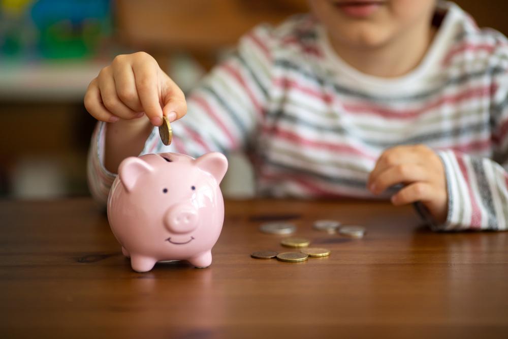 Criança colocando a moeda dentro de um cofre em formato de porco cor de rosa claro. O cofre está sob uma mesa de madeira e nela há mais algumas moedas