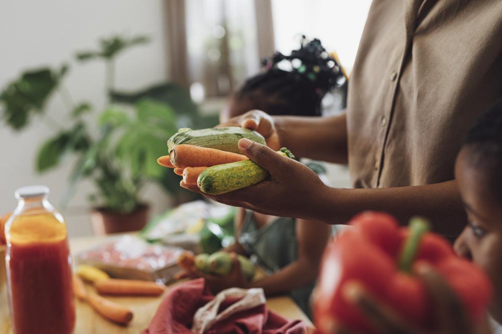 foto de um adulto e duas crianças manuseando vegetais: abobrinhas, cenouras e pimentões, aliemntos que são fundamentais para a alimentação de uma criança vegetariana.