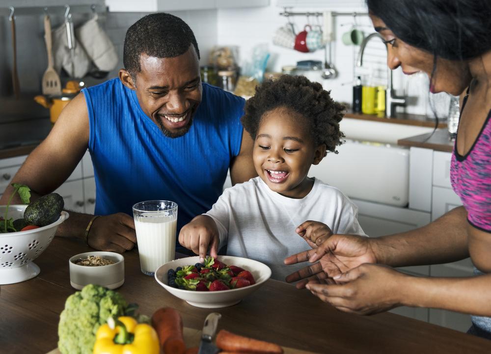 Foto de uma família composta por pai, mãe e criança. Eles estão sorrindo, sentados em frente a uma mesa, onde há refeições saudáveis: morangos, leite e verduras. 