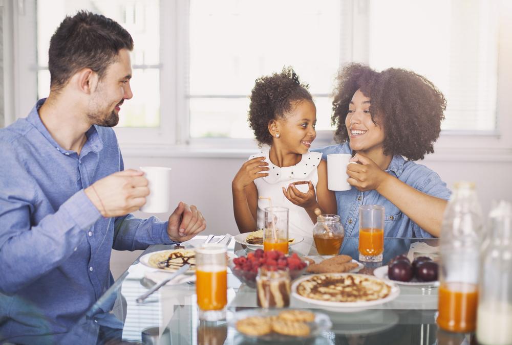 Foto de uma família composta por pai, mãe e criança. Eles estão sorrindo, sentados em frente a uma mesa, onde há um café da manhã nutritivo