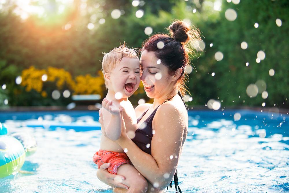 Uma mulher segura uma criança com deficiência no colo, dentro de uma piscina. 