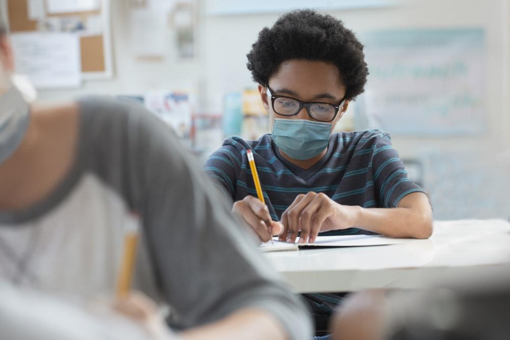 Uma criança negra, vestindo uma camiseta listrada e máscara hospitalar, anota em seu caderno durante a aula. As crianças negras são um dos grupos mais suscetíveis a sofrer racismo na escola, além das crianças indígenas e amarelas.