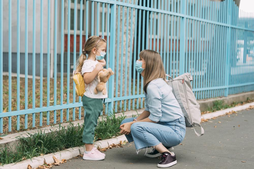 Uma mãe está agachada em frente a sua filha, perto de um portão. A menina está de mochila, olhando para a mãe e segurando um urso de pelúcia.