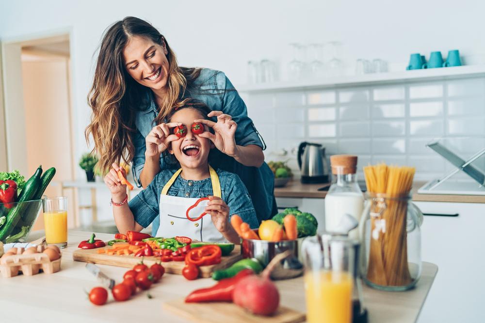 Uma mãe e sua criança estão na cozinha, com diversos alimentos sobre uma bancada. Ela segura dois tomates pequenos na altura dos olhos da criança e ambas estão sorrindo.