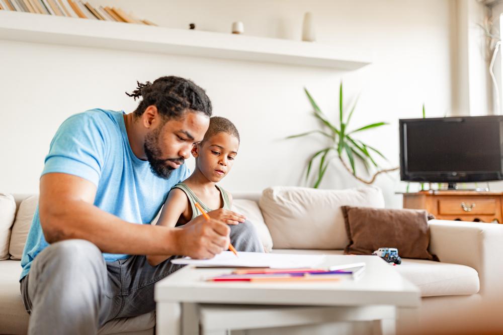 Um pai e seu filho estão em uma sala de estar, desenhando em um papel com lápis de cor.