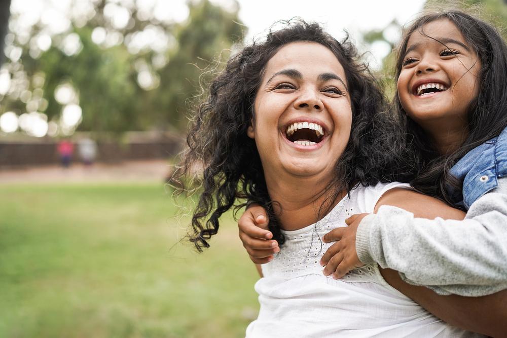 A foto mostra uma criança nas costas das mães sorrindo enquanto estão em um ambiente aberto