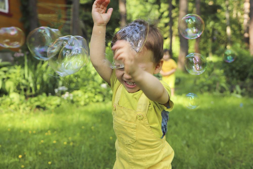 Um menino sorri com as mãos para o alto enquanto passa por bolhas de sabão em um ambiente externo arborizado, com outro menino ao fundo. A foto faz alusão ao tema de brincadeiras ao ar livre. 