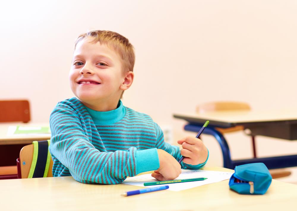 Criança aparentando ter 7 anos de idade, branca, loira, veste camiseta de manga longa azul sentado na classe da escola. Na mesa escolar, materiais para desenho como lápis e papel.