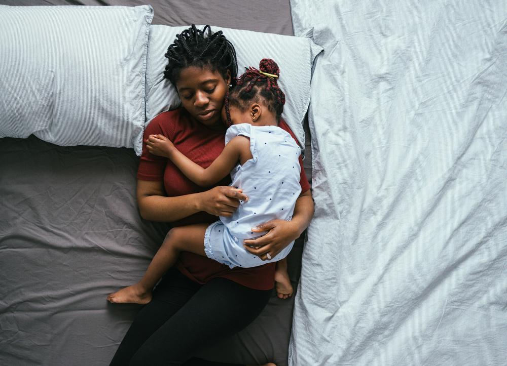Foto de uma mãe solo dormindo com a filha no colo em sua cama. As duas estão no meio da cama que está com as laterais vazias
