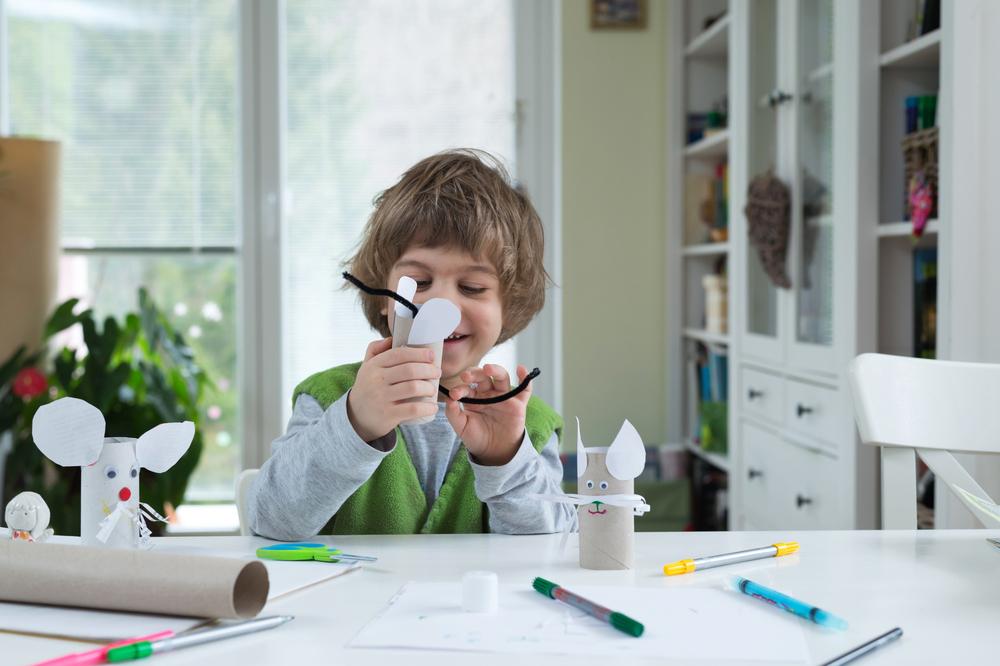 Um menino sorrindo monta brinquedos usando materiais recicláveis espalhados pela mesa de jantar, como folhas de papel, canetas coloridas e rolos de papel higiênico. 