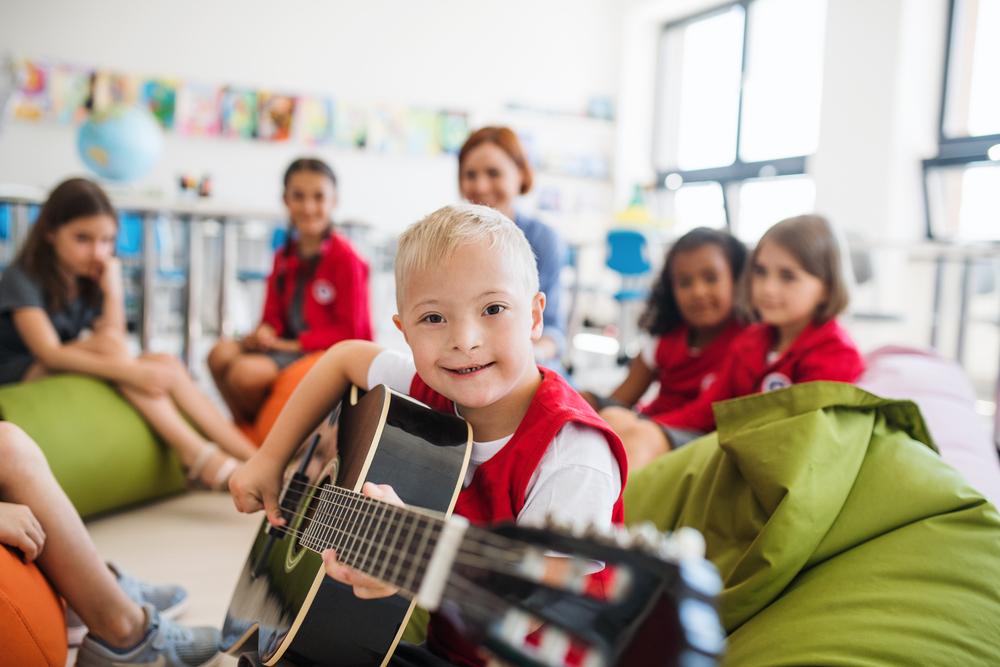 uma criança sorrindo brinca com o violão, ao fundo uma turma de colegas e a professora em sala de aula.